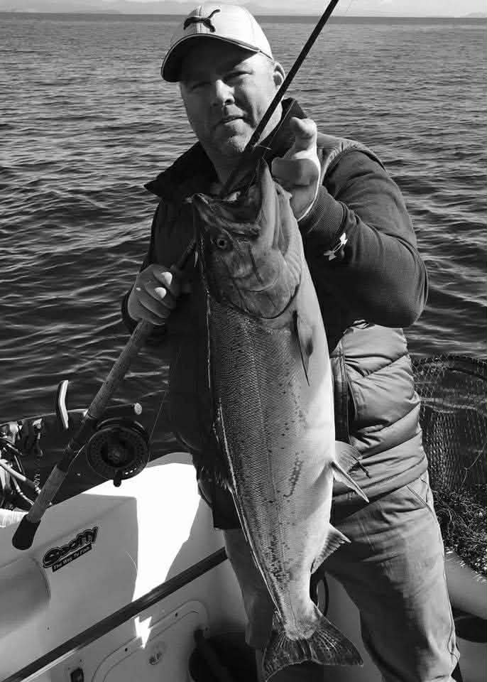 A man holding a large fish he caught, on a boat. The photo is in greyscale, calm waters are visible in the background.